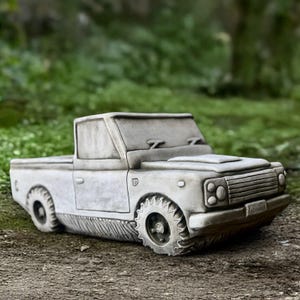 Puede incluir: Escultura de una camioneta pickup de hormigón gris con detalles, incluyendo un capó, faros y una placa de matrícula. La camioneta tiene ruedas grandes y estilizadas y está sobre un fondo verde borroso.