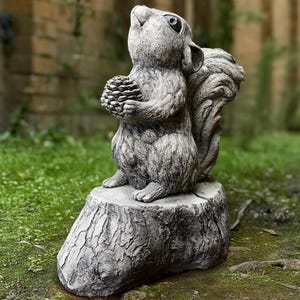 Puede incluir: Una estatua de ardilla de piedra gris que sostiene una piña, de pie sobre una base de piedra gris en forma de tocón de árbol. La ardilla está mirando hacia arriba con la boca abierta.