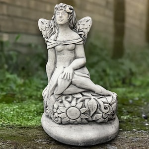 Puede incluir: Una estatua de jardín de piedra de una hada sentada con alas detalladas y una base floral. El hada tiene pelo largo y una expresión serena. La estatua está hecha de piedra gris claro.