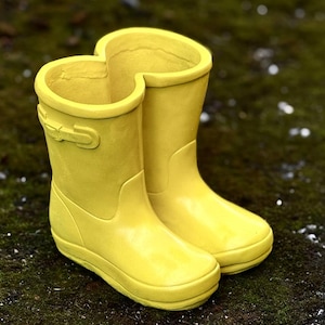 May include: A pair of bright yellow rain boots. The boots are made of a smooth, glossy material and have a rounded toe and a buckle detail on the side. The boots are designed for children and are sitting on a mossy surface.
