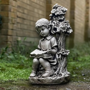 May include: A grey concrete statue of a child reading a book, seated beside a tree with flowers. The statue has a detailed design, suitable for garden or outdoor decor, and is made of concrete.