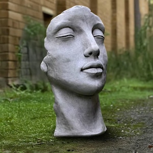 Könnte beinhalten: Eine graue Steinskulptur eines menschlichen Gesichts mit geschlossenen Augen. Die Skulptur ist eine Büste, die Kopf und Hals zeigt. Das Gesicht hat glatte Züge und ist vor dem Hintergrund von grünem Gras und einem Backsteingebäude platziert.