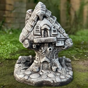 Puede incluir: Una casa de hadas de piedra gris con una base de tronco de árbol y un techo de paja. La casa tiene dos ventanas y una puerta. La base tiene una pequeña puerta y dos pequeñas macetas.