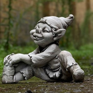 Puede incluir: Una escultura de gnomo de jardín de piedra gris con un sombrero puntiagudo, sentado con una pierna cruzada sobre la otra. El gnomo tiene una expresión traviesa en su rostro.