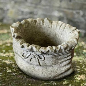 Puede incluir: Una jardinera de piedra gris desgastada con forma de saco atado. El borde superior tiene un diseño con volantes y un lazo decorativo esculpido en la parte delantera. Adecuada para uso en exteriores, añade un toque rústico a los jardines.