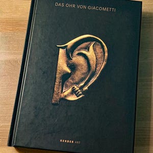 May include: A hardcover book titled "DAS OHR VON GIACOMETTI" with a gold-colored ear illustration on the cover. The book has a black cover with the text "KERBER ART" at the bottom. The book is lying on a wooden surface.