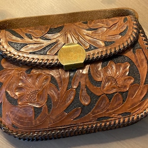 May include: A brown leather bag with a floral design. The bag features a flap closure with a brass clasp. The leather is embossed with flowers and leaves, and the edges are stitched with a braided pattern.
