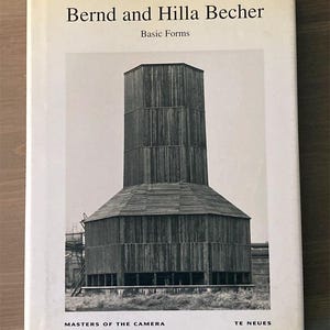 May include: A book titled "Bernd and Hilla Becher Basic Forms" featuring a black and white photograph of a cylindrical industrial structure. The book cover is white with black text and the "Masters of the Camera" and "TE NEUES" printed at the bottom.
