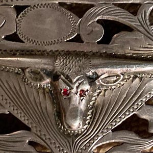 May include: A silver belt buckle with an intricate design featuring a bull's head with red gemstones for eyes. The buckle is decorated with floral patterns and a filigree design.