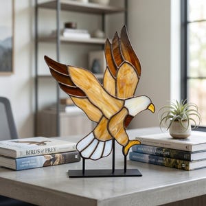 Puede incluir: Una escultura de águila de vidrieras con las alas extendidas, con tonos marrones, ámbar y blanco. El águila está montada sobre un soporte de metal negro. La escultura se exhibe sobre una superficie gris, con libros y una planta en el fondo.