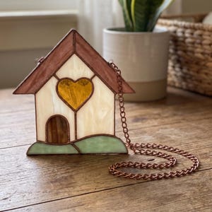 May include: A house-shaped stained glass suncatcher featuring a cream-coloured body, a brown roof, a green base, and a central yellow heart-shaped window. The piece is framed in copper-coloured metal and includes a matching copper-coloured metal chain.