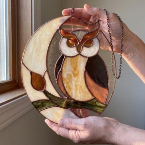 May include: A circular stained glass suncatcher featuring an owl perched on a branch against a crescent moon background. The piece is constructed with various shades of amber, brown, cream, and green glass, held together by dark metal soldering.