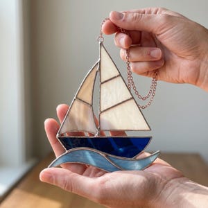 Puede incluir: Un atrapasoles de vitral con forma de velero, con velas color blanco hueso, un casco azul oscuro y olas azul claro. Las piezas de vidrio están unidas por líneas de soldadura de metal color cobre. Incluye una cadena metálica a juego.