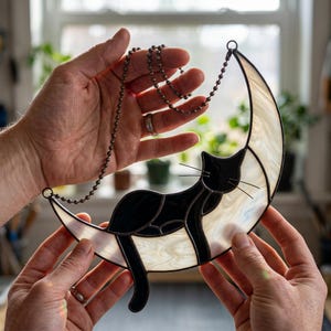 Puede incluir: Un gato de vidriera descansa sobre una luna creciente. El gato es negro con detalles blancos, y la luna es blanca con brillo nacarado. Una cadena de metal oscuro está unida para colgar.