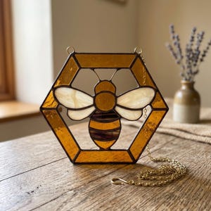 May include: A hexagonal stained glass suncatcher featuring a central bee design with amber-coloured glass panels, white wings, and dark brown stripes. The piece is framed in dark metal and includes a gold-toned metal chain for hanging purposes.