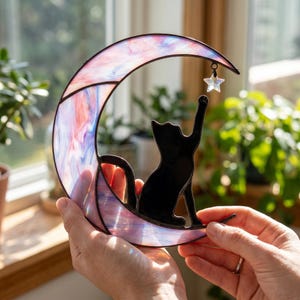 Op de afbeelding: Een glas-in-lood halve maan in tinten roze, blauw en wit, met het silhouet van een zwarte kat die op de onderste boog zit en reikt naar een kleine, heldere, stervormige glazen bedel die aan de bovenkant van het maanframe hangt.
