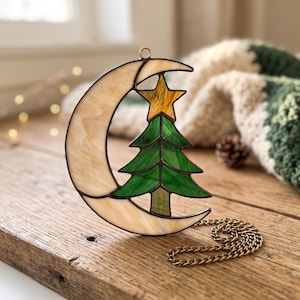 May include: A stained glass suncatcher featuring a crescent moon in cream-colored marbled glass, a green glass Christmas tree, and a yellow glass star at the top. The piece is framed in dark metal solder and includes a matching metal hanging chain.