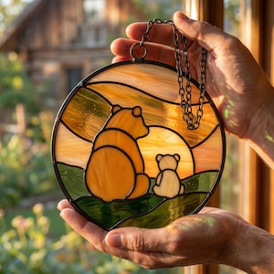 Könnte beinhalten: Ein runder Buntglas-Sonnenfänger mit einem großen und einem kleinen Bären, die zusammen auf einer grünen Wiese sitzen und in einen goldenen Sonnenuntergang blicken. Das Stück ist schwarz eingefasst und hat eine Metallkette zum Aufhängen.