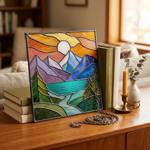 May include: A square stained glass panel featuring a landscape with a white sun, blue and purple mountains, a turquoise lake, and green pine trees. The piece is framed in dark metal and includes a hanging chain attached to the bottom right corner.