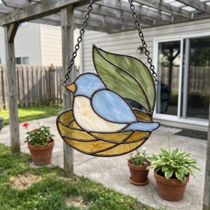 Op de afbeelding: Een glas-in-lood zonnevanger met een blauwe en crèmekleurige vogel in een geel nest, geplaatst voor een groot groen blad. Het stuk is omlijst met donkere metalen soldeerlijnen en hangt aan een zwarte metalen ketting om op te hangen.