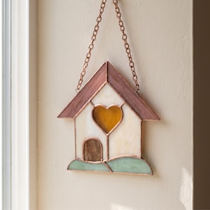 Könnte beinhalten: Ein Buntglas-Hausornament mit kupferfarbenem Dach, cremefarbenen Wänden, einem zentralen bernsteinfarbenen herzförmigen Fenster, einer braunen Bogentür und einem hellgrünen Sockel. Das Stück ist an einer Kupferkette zum Aufhängen befestigt.