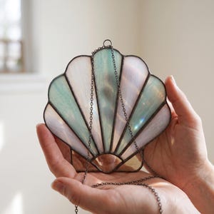 May include: A stained glass seashell suncatcher featuring iridescent white and pale blue glass panels held together by dark metal solder lines. The shell-shaped piece includes a thin metal chain attached to the top for hanging purposes.