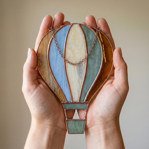 Peut inclure: Un attrape-soleil en vitrail en forme de montgolfière avec des panneaux verticaux dans des tons ambre, bleu clair, crème et vert sauge. Les pièces de verre sont assemblées par des lignes de soudure en cuivre et incluent une chaînette.