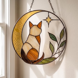 May include: A circular stained glass suncatcher featuring a cat sitting in front of a yellow crescent moon. The design includes a small yellow star and green leaves, all framed in dark metal with a chain for hanging against a light wall.