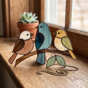 Puede incluir: Un atrapasoles de vidrieras con tres pájaros posados en una rama. El pájaro de la izquierda es blanco y marrón, el central es azul claro y azul oscuro, y el de la derecha es amarillo y verde. Una cadena de metal está unida a la rama.