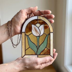 May include: A stained glass suncatcher featuring an arched window design with a central white tulip flower and green leaves. The background consists of amber-colored glass panels framed in dark metal, with a thin metal chain attached for hanging.