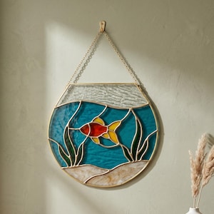 May include: Stained glass wall hanging featuring a goldfish in a bowl design. The fish is red and yellow, set against a turquoise blue background with green seaweed and a sandy base. The piece is framed in gold and hangs from a chain.