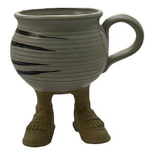 Puede incluir: Una taza de cerámica blanca con rayas negras y tres patas con forma de zapatos. La taza tiene un asa y está hecha en un estilo rústico.