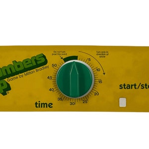 Op de afbeelding: Een gele timer met een groene knop voor het Numbers Up spel van Milton Bradley. De timer heeft een wijzerplaat met nummers van 5 tot 50 en de woorden "Niet voorbij dit punt draaien" en "Alleen in de richting van de pijl draaien". De timer heeft ook een start/stop-knop.