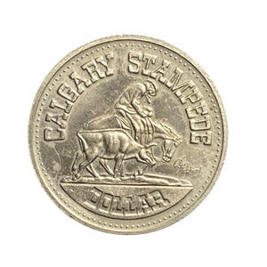 May include: A silver Calgary Stampede dollar coin. The coin features a cowboy riding a horse and a steer. The words "CALGARY STAMPEDE" and "DOLLAR" are inscribed around the edges.