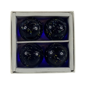 May include: Four dark blue marbles with colourful speckles are displayed in a divided white cardboard box. The marbles have a glossy finish and are arranged in two rows of two.