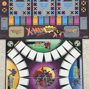 Op de afbeelding: Twee spelborden, een voor het spel Uncanny X-Men Alert! en een voor het spel Spawn: The Game. Het X-Men-bord heeft een zwarte achtergrond met gele, blauwe en paarse accenten. Het Spawn-bord heeft een paarse en zwarte achtergrond met een rood en wit Spawn-personage in het midden.