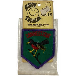 Puede incluir: Un parche de planchado vintage Happy Traveler con un fondo verde y un borde morado. El parche representa un petirrojo posado en una rama con la palabra "CONNECTICUT" en letras amarillas. El parche está empaquetado en una bolsa de plástico transparente con una tarjeta amarilla que dice "Happy Traveler" con una cara sonriente y "Stuckey's Souvenir Emblem 129". La tarjeta también dice "Coselos en gorras, chaquetas, camisas, etc."