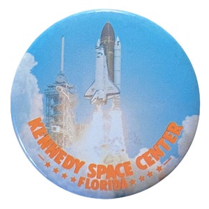 May include: A round, blue button with a picture of a space shuttle launching from the Kennedy Space Center in Florida. The text "KENNEDY SPACE CENTER - FLORIDA" is printed in orange around the edge of the button.