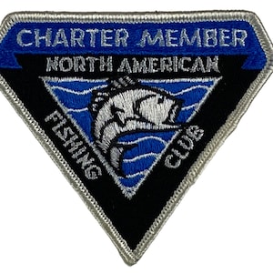 May include: A silver and blue embroidered patch with the text "Charter Member North American Fishing Club". The patch features a white fish jumping out of blue water.