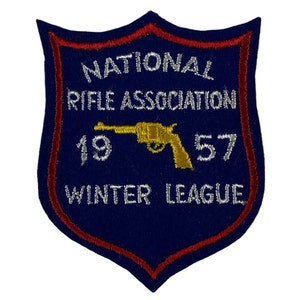 May include: A navy blue shield-shaped patch with red trim and white embroidered text. The text reads "NATIONAL RIFLE ASSOCIATION 1957 WINTER LEAGUE" with a gold pistol graphic.