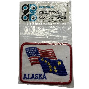 以下が含まれることがあります： アメリカの旗とアラスカ州の旗が刺繍された白いパッチで、旗の下に「ALASKA」という文字が書かれています。パッチは、透明なビニール袋に包装されており、「Embroidered by Goldenland Crafts & Crafts」と書かれたラベルが付いています。