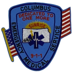 May include: Blue and white embroidered patch with the text "Columbus Emergency Medical Service" and "Dedicated to One More" with a sunrise and a star of life graphic. The patch also features a small American flag on each side.