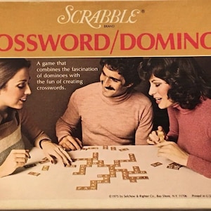 Pode incluir: Um jogo de tabuleiro chamado Scrabble Crossword/Dominoes. A caixa mostra três pessoas jogando o jogo com peças de dominó que têm letras. O jogo combina a diversão de criar palavras cruzadas com a fascinação dos dominós.
