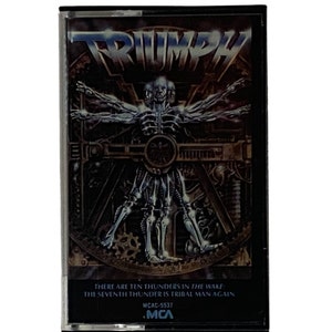 Op de afbeelding: Een cassettebandje met een zilveren en zwarte hoes met een robotfiguur met uitgestrekte armen. De tekst "TRIUMPH" is in grote, vetgedrukte letters bovenaan de hoes gedrukt. De tekst "THERE ARE TEN THUNDERS IN THE WAKE THE SEVENTH THUNDER IS TRIBAL MAN AGAIN" is onderaan de hoes gedrukt. De tekst "MCAC-5537 MCA" is rechtsonder gedrukt.