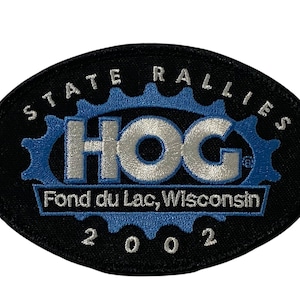 Puede incluir: Parche ovalado negro con texto blanco que dice "State Rallies HOG Fond du Lac, Wisconsin 2002". El logotipo de HOG es plateado con un engranaje azul detrás.