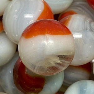 May include: A close-up of a collection of white and orange glass marbles. The marbles have a swirling pattern and are translucent.