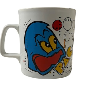 May include: White ceramic mug with a blue cartoon character with a red tongue and yellow eyes. The character is surrounded by colorful Pac-Man-like shapes and the word "PAC-MAN" is printed on the mug.