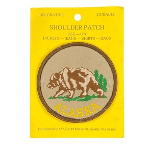 May include: A round shoulder patch featuring a brown and white bear walking on green grass, with the word "ALASKA" in yellow. The patch is on a yellow backing with the words "DECORATIVE" and "DURABLE".
