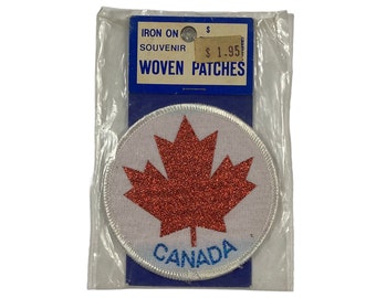 Puede incluir: Un parche tejido redondo blanco para planchar con una hoja de arce roja con purpurina y la palabra "CANADA" en letras azules. El parche está empaquetado en una bolsa de plástico transparente con una etiqueta de precio azul y blanca que dice "IRON ON SOUVENIR WOVEN PATCHES $1.95".