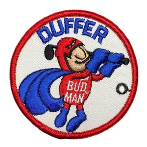 Puede incluir: Parche bordado con un personaje de superhéroe de dibujos animados en rojo y azul, sosteniendo un objeto azul. El parche tiene la palabra "DUFFER" en azul y "BUD MAN" en rojo, en un diseño circular.
